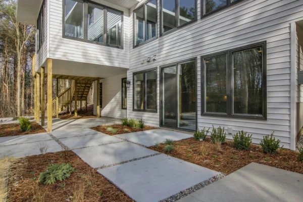 IMG_6390Blended Modern house with large windows, white siding, and a paved walkway surrounded by small plants and trees.