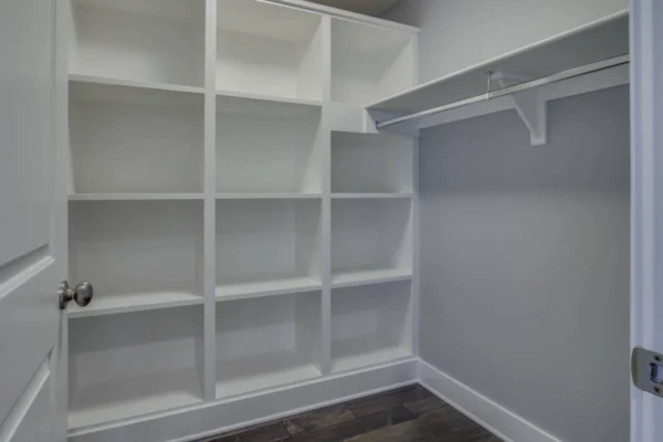 IMG_6486Blended Empty walk-in closet with white shelves and hanging rod on dark wooden floor.