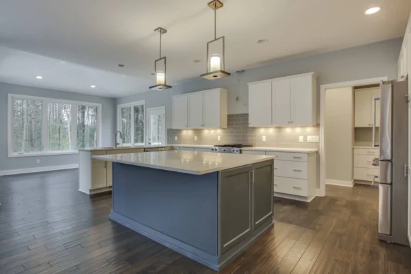IMG_6561Blended Modern kitchen with a gray island, dark wood floors, pendant lights, and large windows overlooking a wooded area.