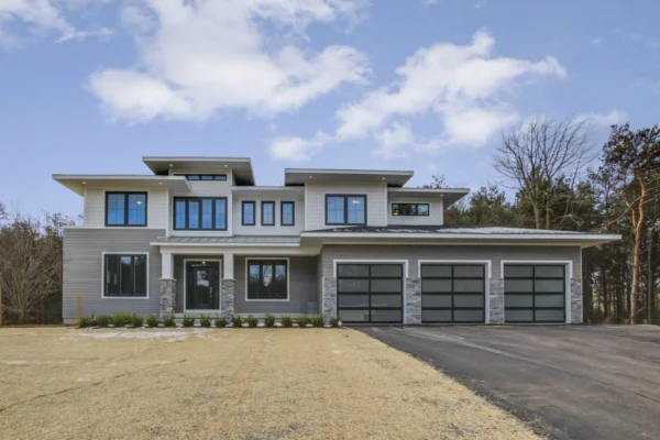 IMG_6588Blended Modern two-story house with large windows, three garage doors, and a paved driveway, set against a cloudy sky.