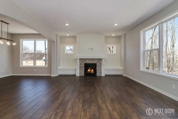 IMG_7661_blended Spacious living room with a lit fireplace, wooden floor, large windows, and modern light fixtures.