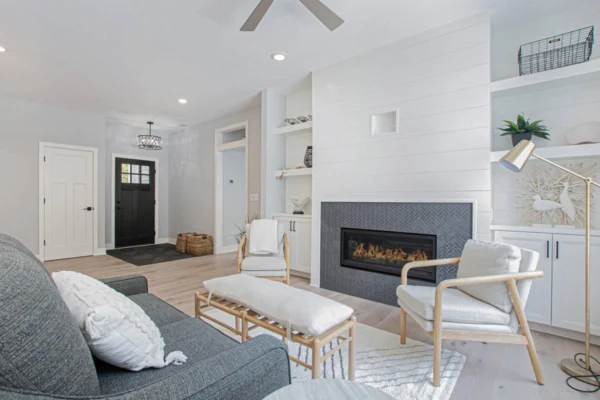 IMG_8458-1 – Copy Modern living room with gray sofa, armchairs, shiplap wall, sleek fireplace, and open shelves. White and beige decor.