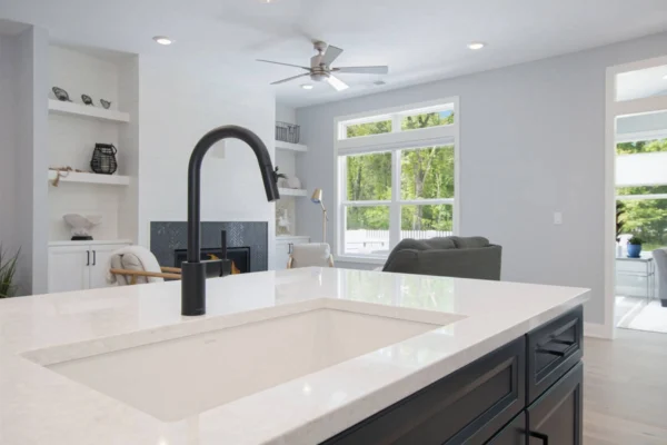 IMG_8473-1 Modern kitchen with black faucet, white countertop, and living area in background with large windows and greenery outside.