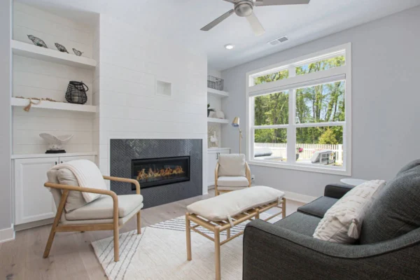 IMG_8482-1 Modern living room with a fireplace, wooden chairs, a gray sofa, and large window overlooking a garden.