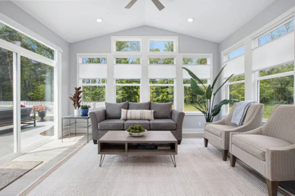 IMG_8490 – Copy Bright sunroom with a gray sofa, two beige chairs, plants, and a coffee table on a light rug, surrounded by windows.