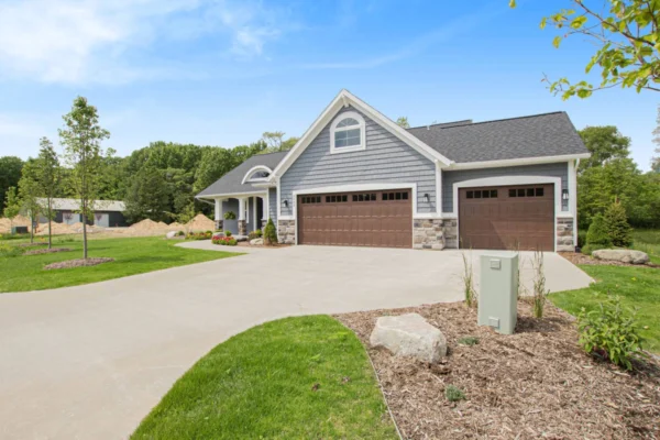 IMG_8525 Suburban house with a three-car garage, gray siding, and stone accents, surrounded by trees and a green lawn.