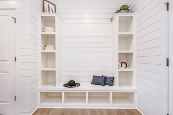 IMG_9940 White mudroom with open shelves, bench, hats, and cushions against shiplap walls.