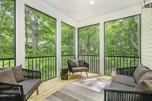 IMG_9946 Screened porch with wicker furniture, cushions, and a rug, overlooking a lush green forest.