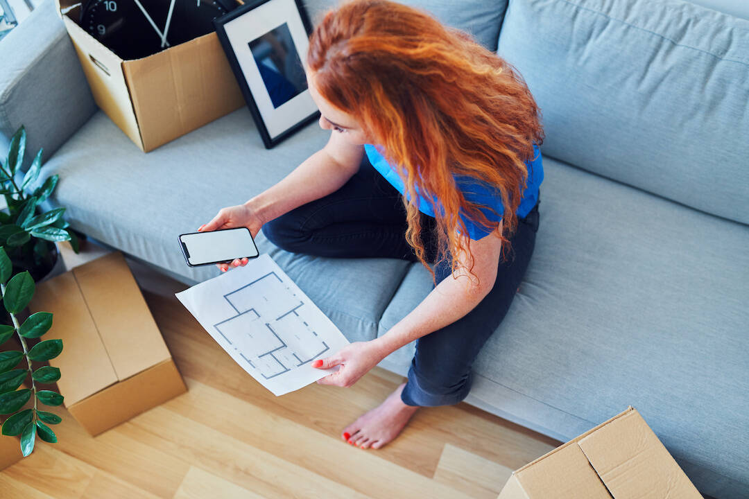 Top view of young woman sitting on sofa with apartment plan and phone